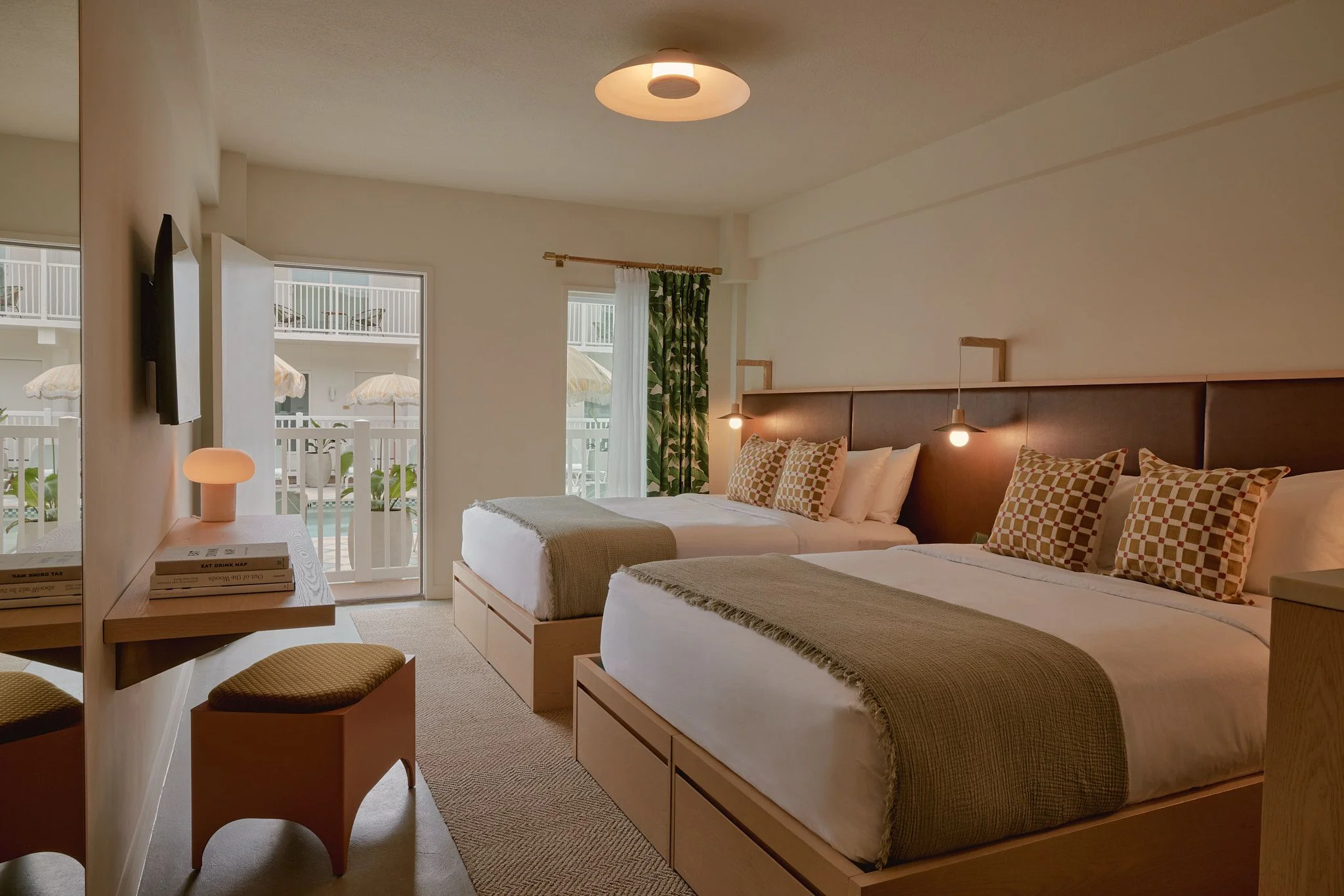 A bright, contemporary hotel bedroom featuring two queen beds with upholstered headboards, warm earth-tone bedding with patterned pillows, and natural wood furnishings, accented by pendant lighting and a circular ceiling fixture; floor-to-ceiling glass doors open to a beachfront balcony with umbrellas and lounge chairs visible outside.