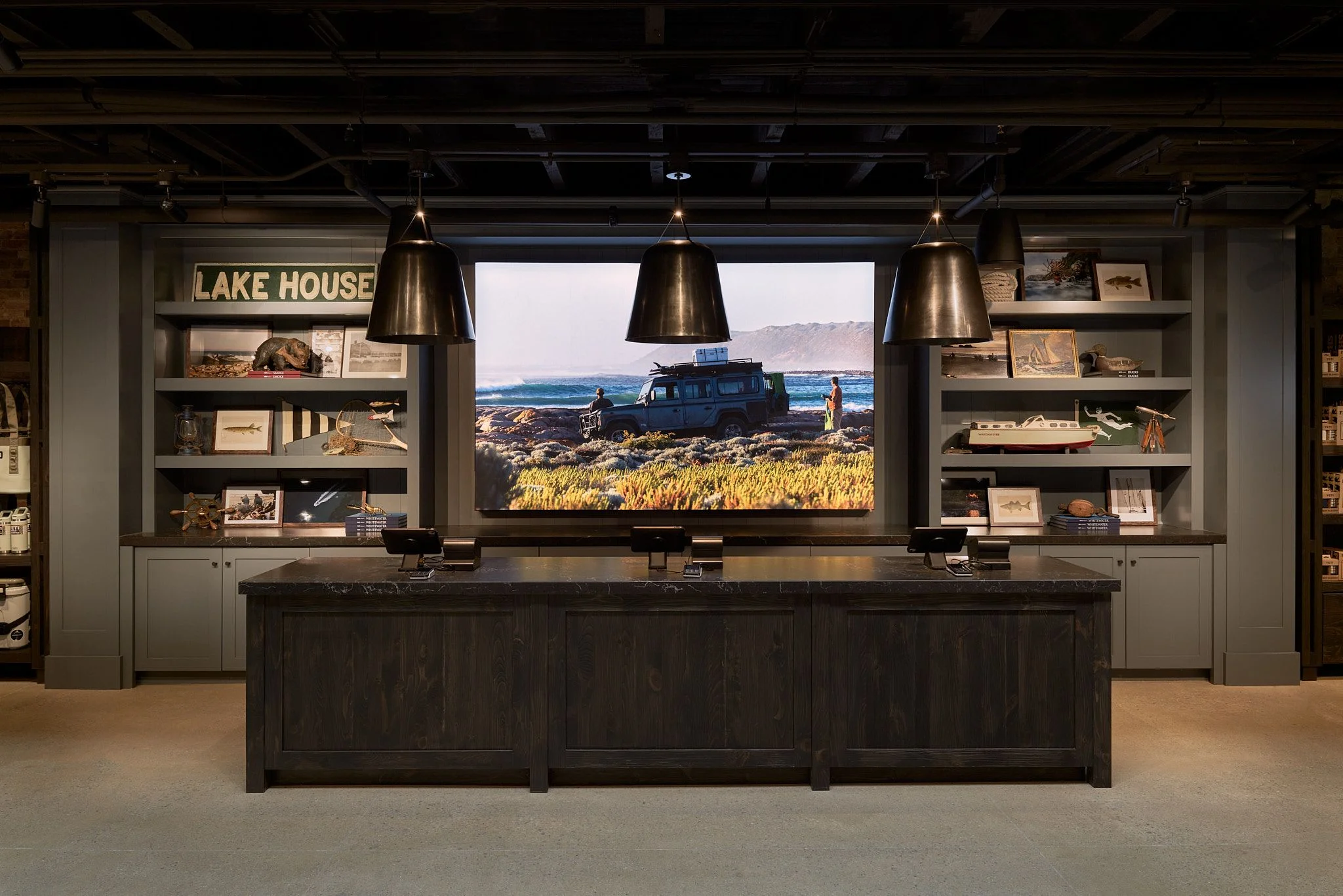 A modern lakehouse interior featuring a dark wood counter and shelving display with vintage photographs and memorabilia, centered around a large landscape photograph of a blue van parked in a golden wheat field overlooking water; three brass pendant lights hang above, and a "Lake House" sign is visible on the left shelving unit.