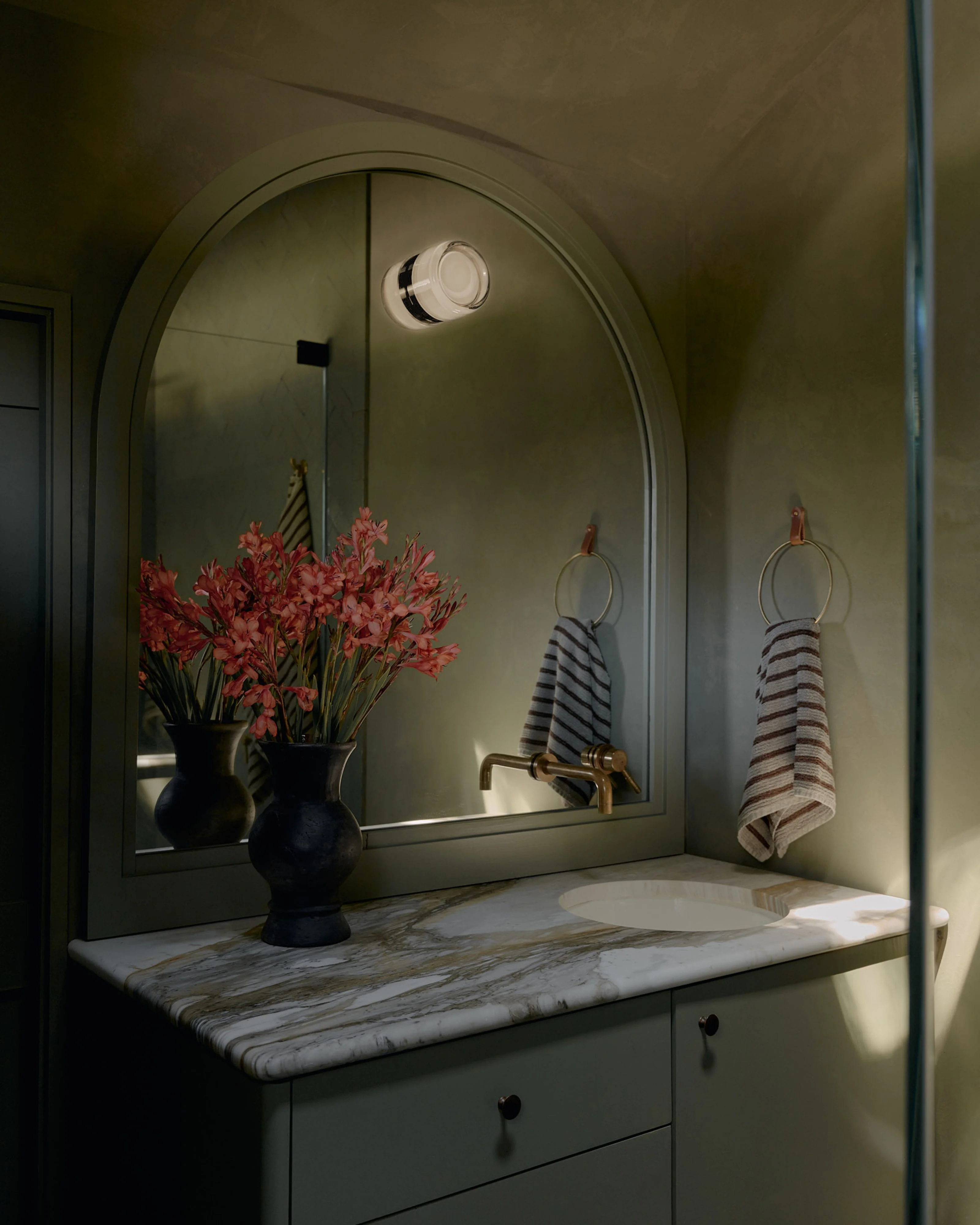 A serene bathroom vanity features a large arched mirror framed in soft sage green, reflecting black vases of vibrant red flowers and striped hand towels hung on brass rings. The marble countertop is paired with a muted green cabinet base, warm sconce lighting, and minimalist modern design elements.
