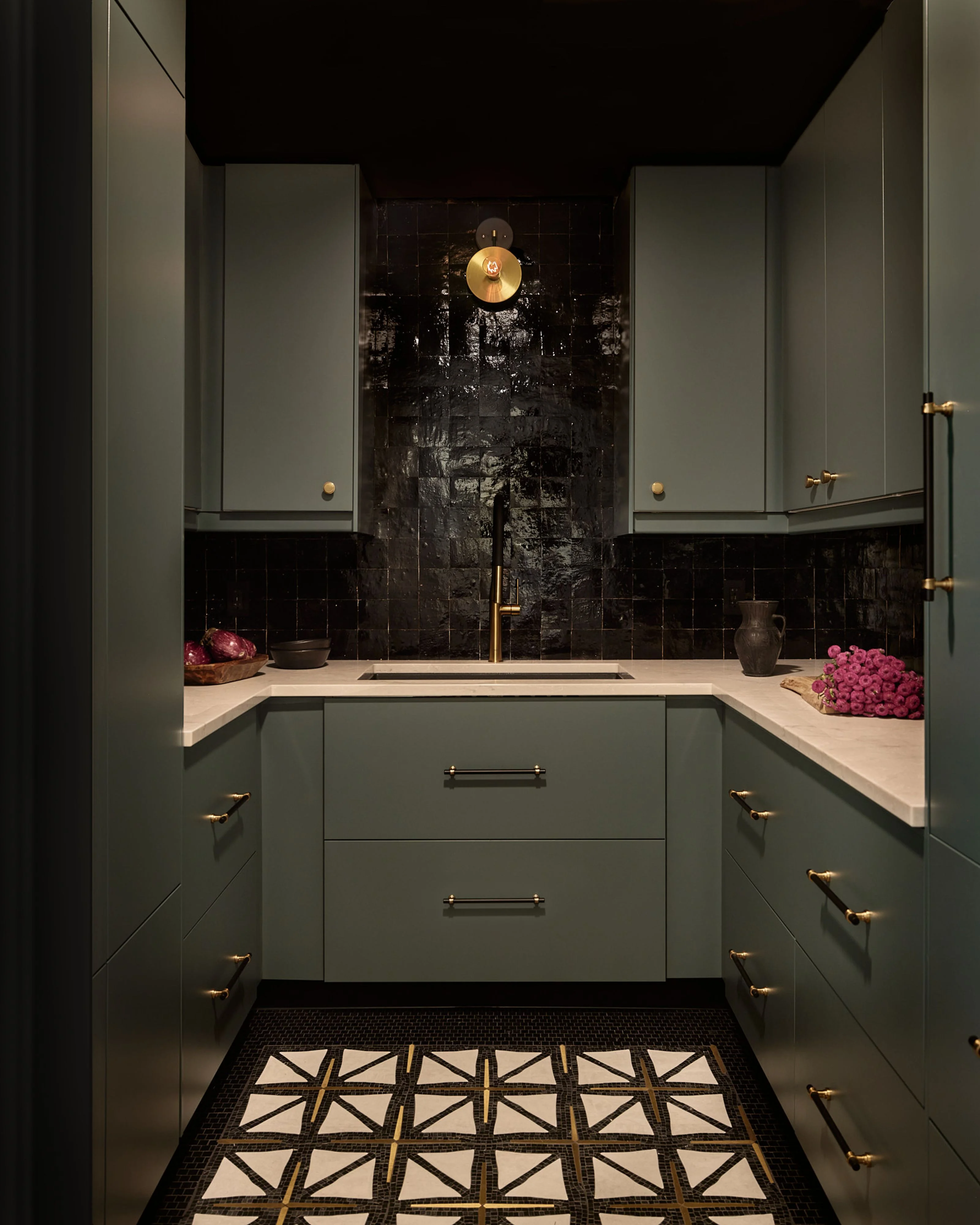 A contemporary kitchen with sage green cabinetry and brass hardware features a dramatic black subway tile backsplash, light stone countertops, and a central sink beneath a brass pendant light. The moody, sophisticated space is accented with pink floral arrangements and a geometric black-and-white patterned floor runner.