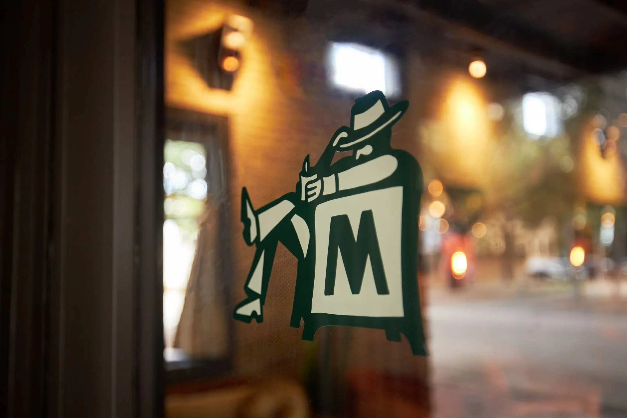 A stylized illustration of a cowboy character wearing a hat and holding a sign with "M" is displayed on a glass storefront window at night, with warm amber interior lighting and blurred street lights visible in the background.