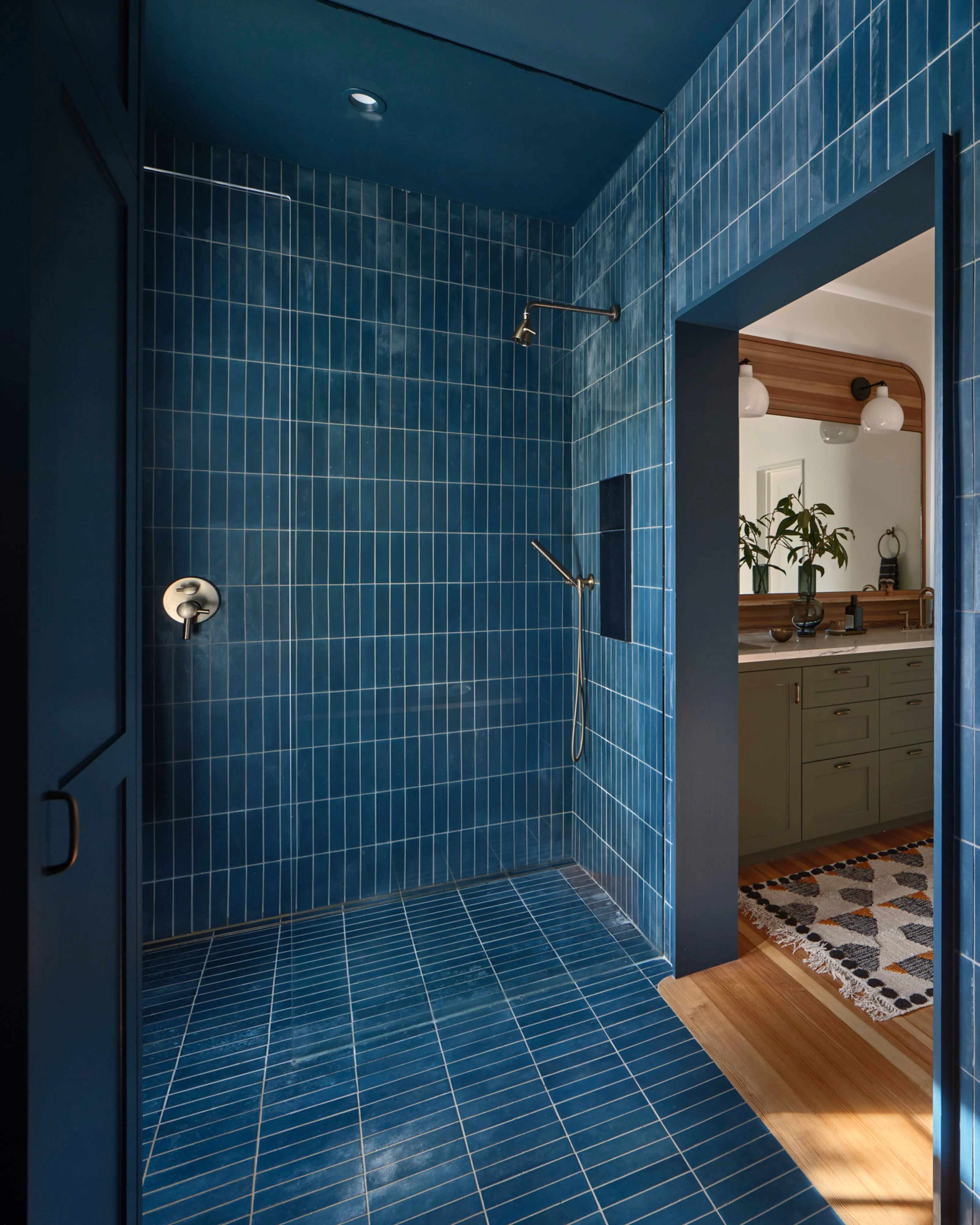 A modern bathroom shower enclosure features deep teal blue vertical tile work on all walls and ceiling, with matching linear floor tiles, complemented by brushed bronze fixtures and a frameless glass partition. The open doorway reveals an adjacent vanity area with olive-green cabinetry and warm wood accents.