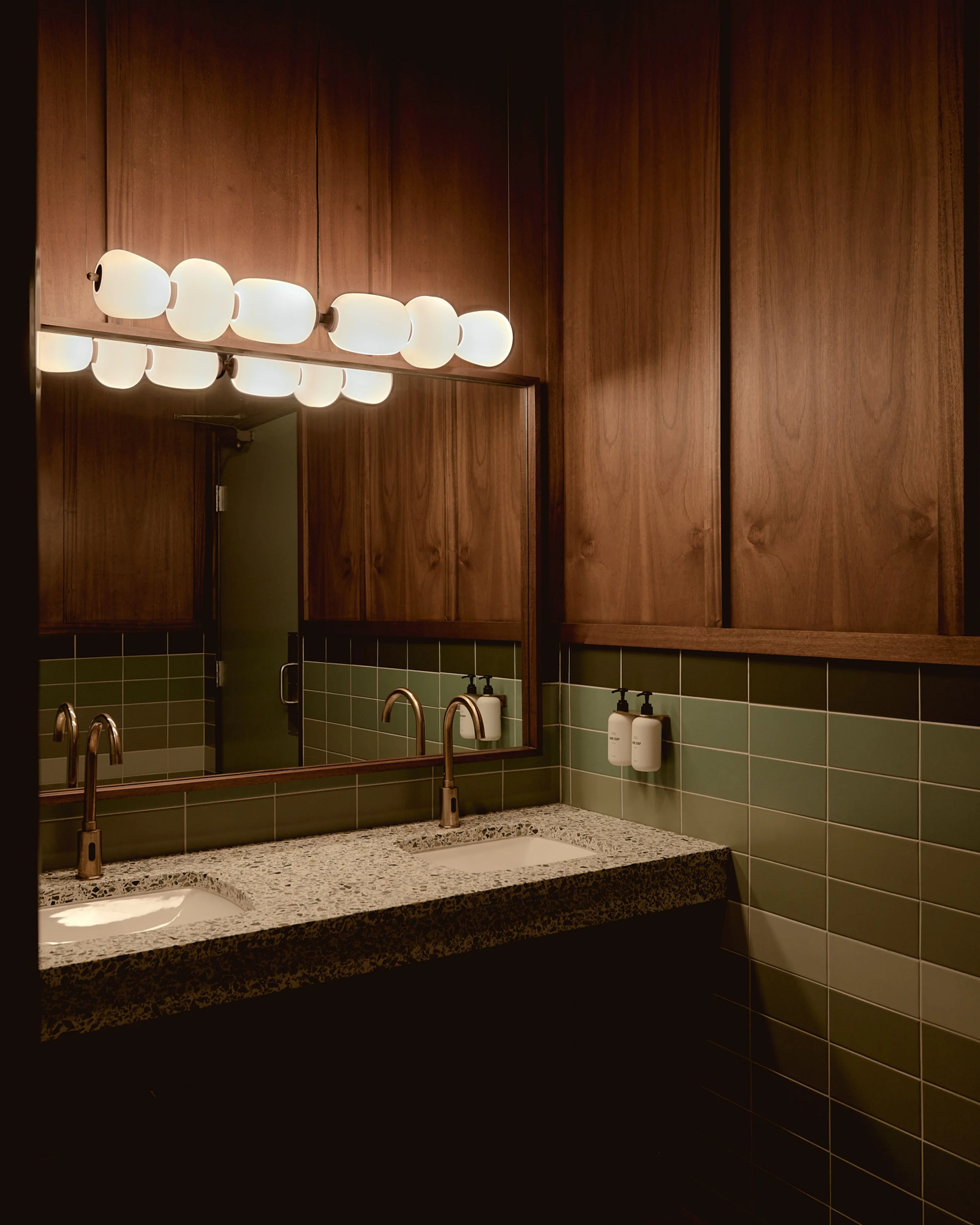 A mid-century modern bathroom features warm wood paneling walls, sage green subway tile wainscoting, and a terrazzo countertop with an integrated sink, illuminated by a sculptural globe vanity light fixture with brass accents.