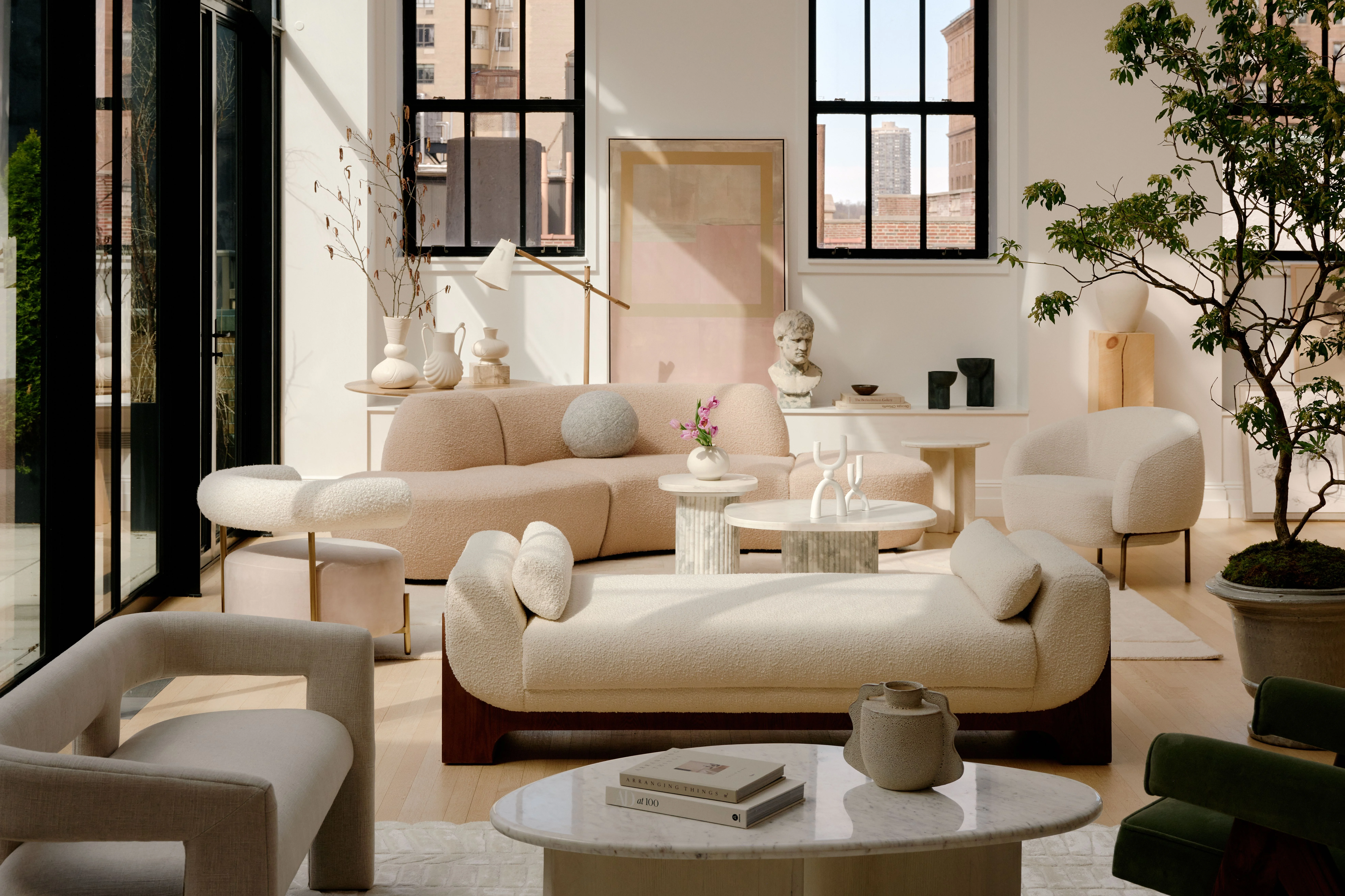A minimalist modern living room features sculptural cream-colored upholstered furniture with curved forms, including a distinctive rounded sofa and organic-shaped chairs arranged around marble and wood coffee tables. Floor-to-ceiling black-framed windows flood the space with natural light, complemented by potted greenery, neutral décor accessories, and a serene palette of whites, beiges, and soft pastels.