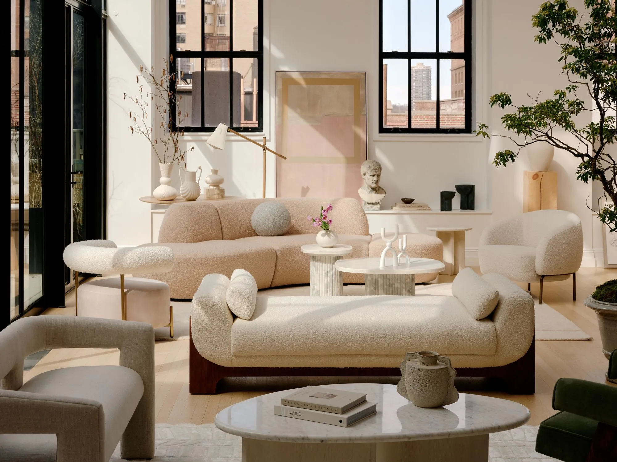 A minimalist modern living room features sculptural cream-colored upholstered furniture with curved forms, including a distinctive rounded sofa and organic-shaped chairs arranged around marble and wood coffee tables. Floor-to-ceiling black-framed windows flood the space with natural light, complemented by potted greenery, neutral décor accessories, and a serene palette of whites, beiges, and soft pastels.