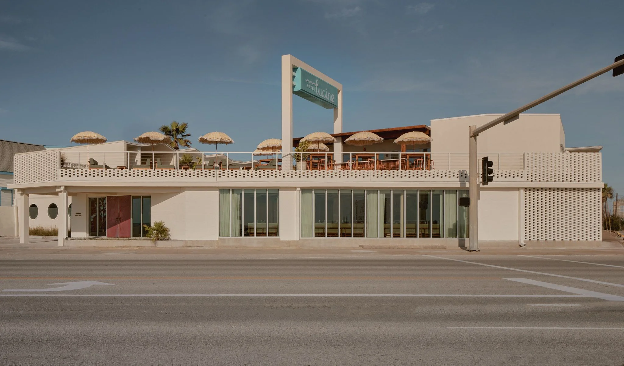 A modernist beachfront restaurant features clean white cubic architecture with geometric perforated screens, expansive glazing, and a rooftop terrace lined with natural umbrellas and palm trees under clear blue skies.