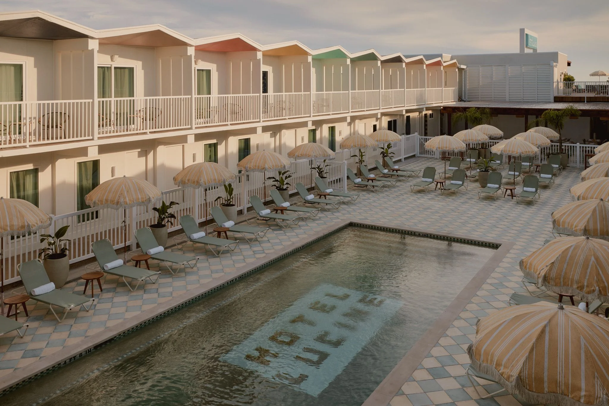 A seaside resort courtyard features a rectangular pool surrounded by a geometric patterned deck with rows of striped umbrellas and lounge chairs, flanked by multi-story cream-colored buildings with peaked rooflines and white railings in a contemporary coastal architectural style.