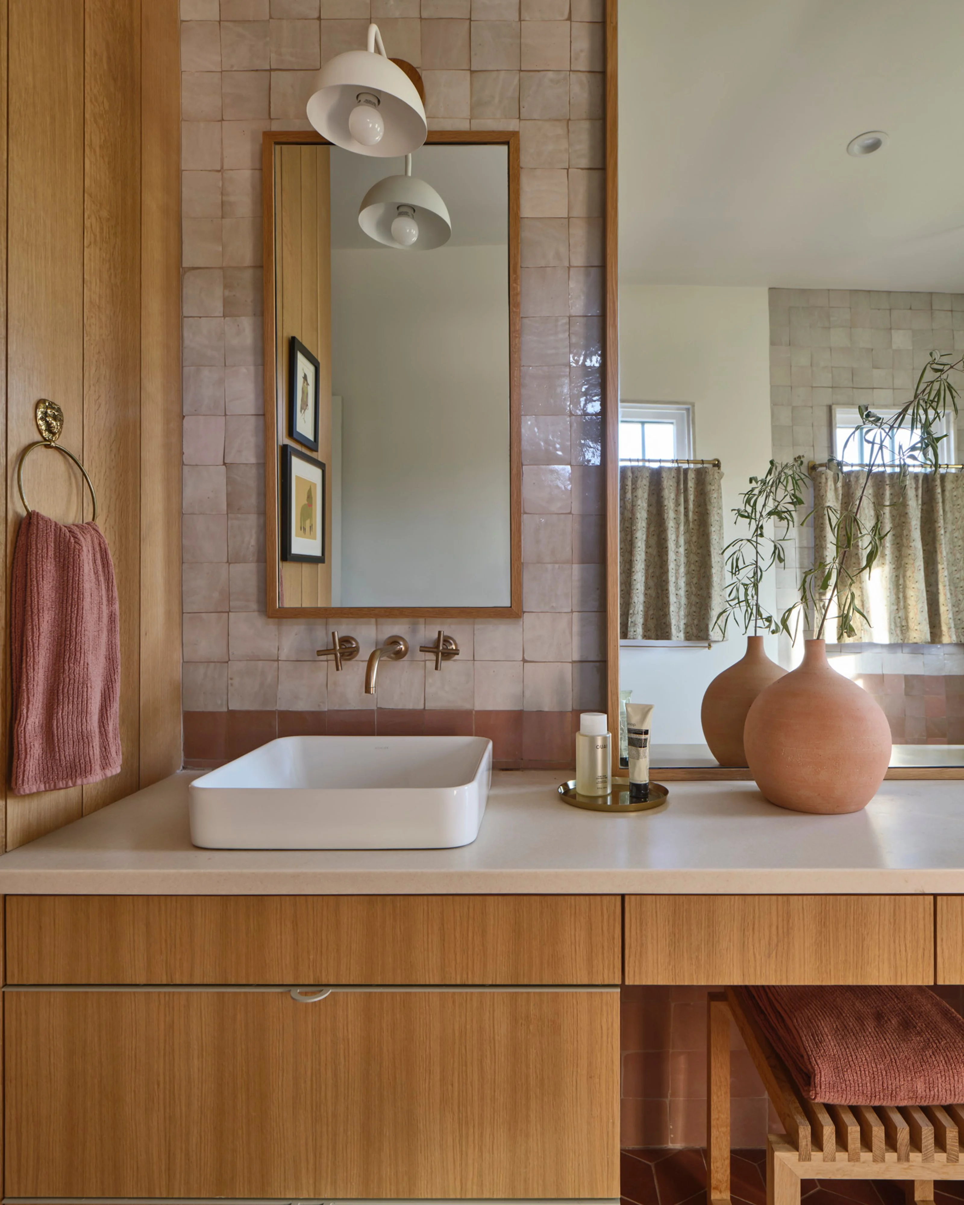 A mid-century modern bathroom features warm wood cabinetry and wall paneling paired with dusty rose tile, centered by a white rectangular vessel sink beneath a wood-framed mirror and dual pendant lights. The serene space is accessorized with a terracotta ceramic vessel, brass fixtures, a burgundy towel, and potted greenery.