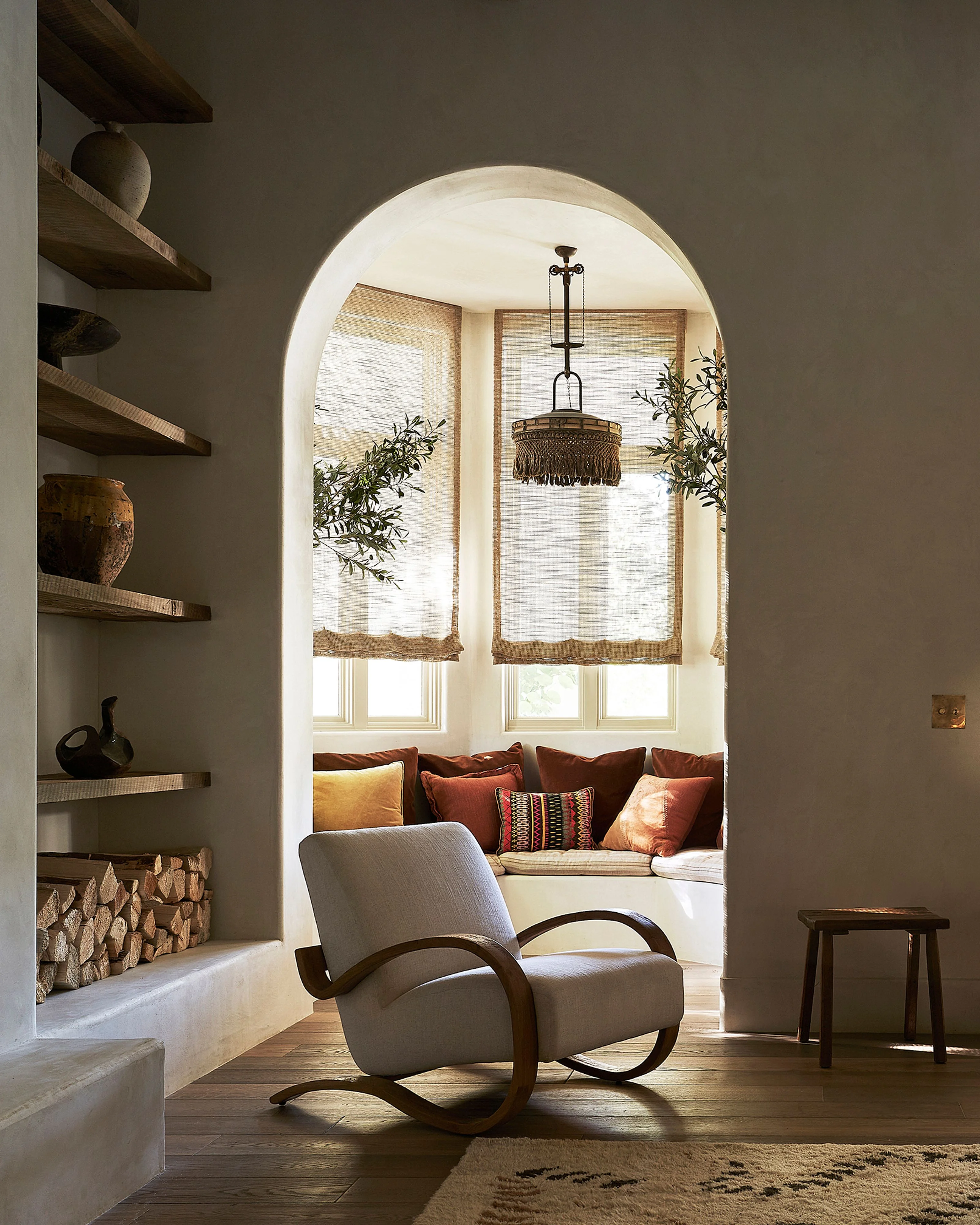 A serene modern interior features a curved archway framing a sunlit window nook with a cushioned bench and woven roman shades, while a sculptural curved armchair in light gray upholstery sits in the foreground beside floating wooden shelves displaying decorative objects and a low firewood storage area.