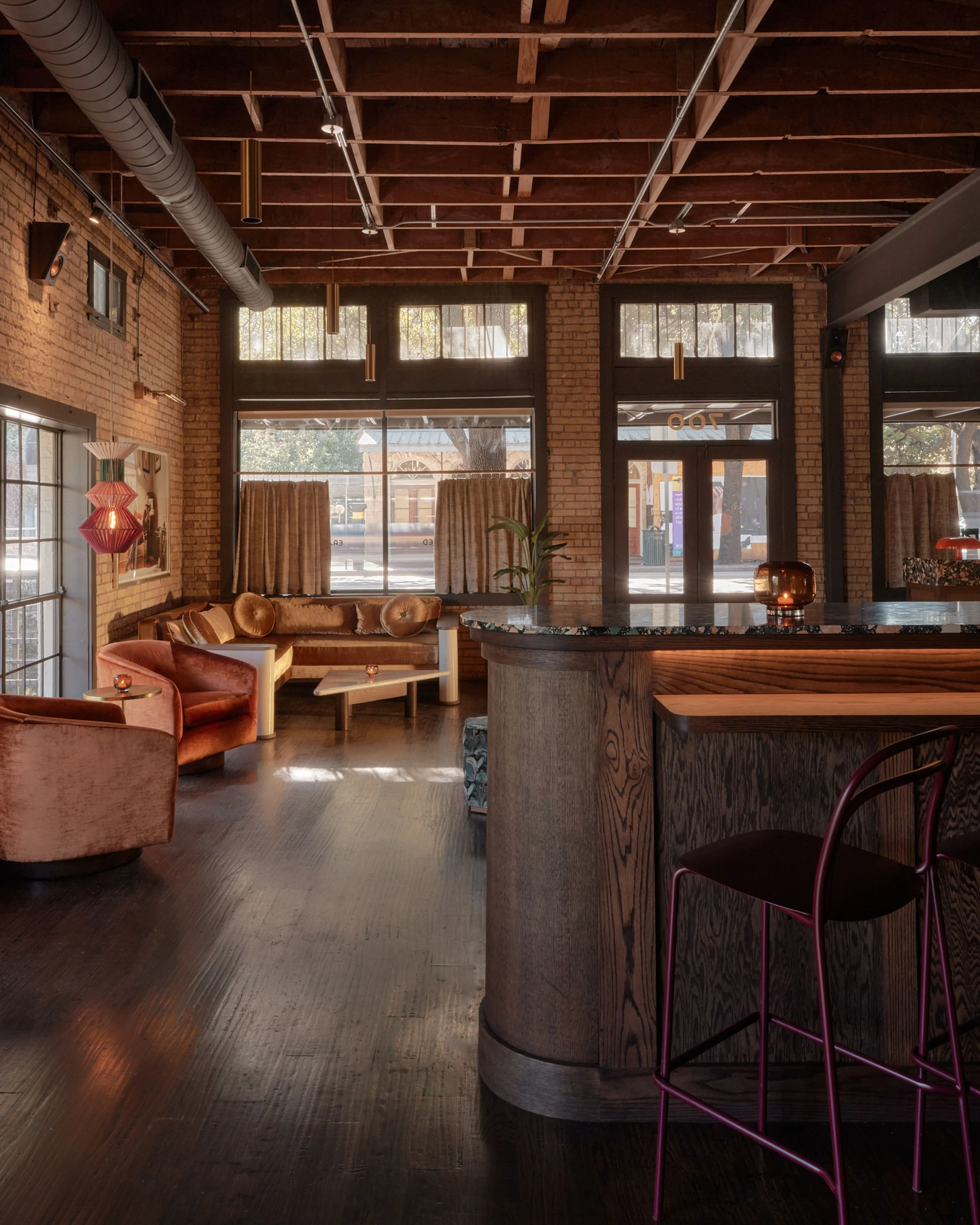 An industrial-style lounge and bar space featuring exposed wooden ceiling beams, red brick walls, and dark hardwood flooring, with warm terracotta and rust-toned upholstered seating arranged around a wooden bar counter. Large windows with earth-toned curtains overlook a street-level storefront, while brass pendant lights enhance the sophisticated, mid-century modern aesthetic.