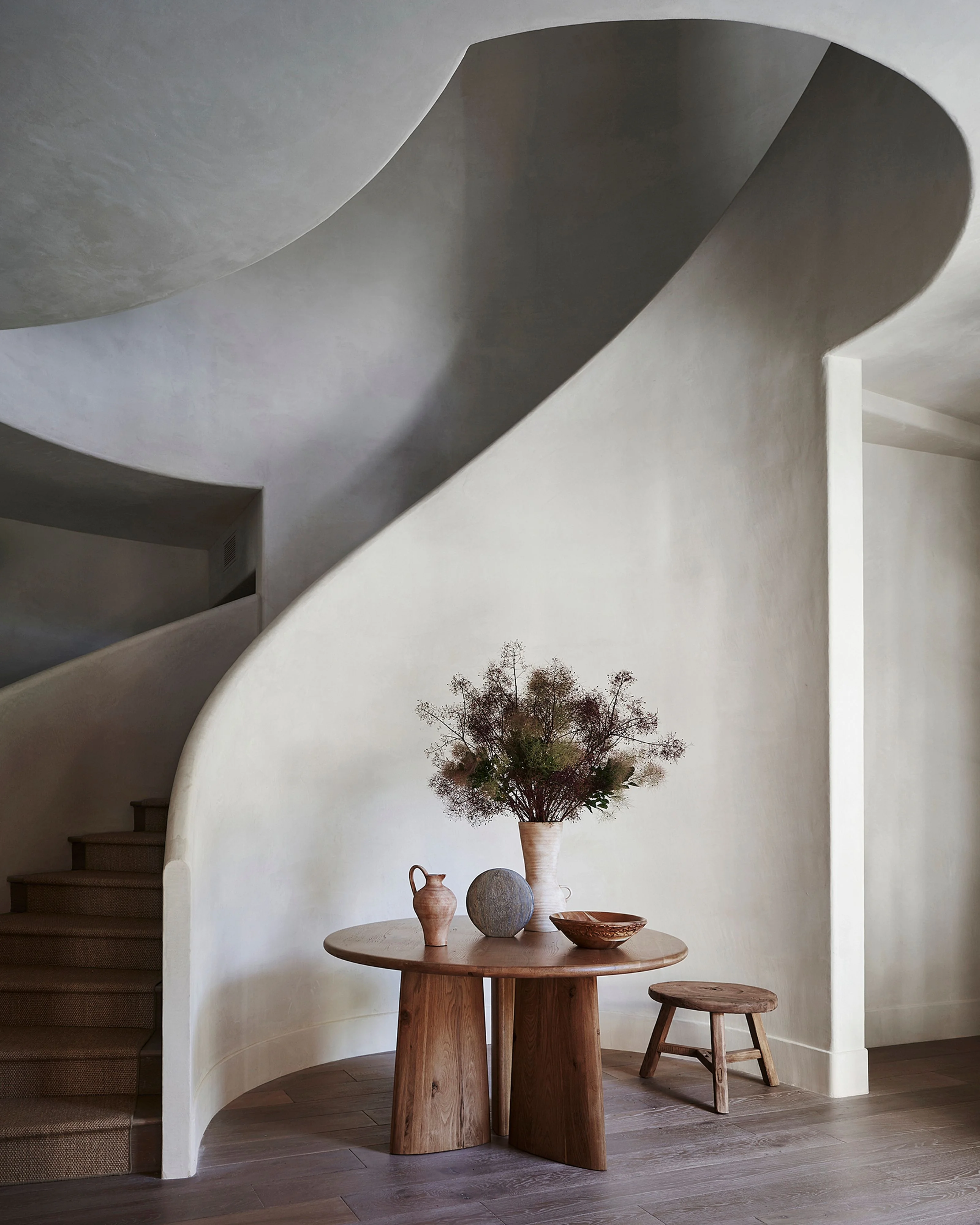 A minimalist interior space featuring a sculptural curved white plaster wall that flows organically around a wooden round table styled with earthy ceramics and dried botanicals, with wooden steps visible on the left and a small wooden stool beside the table, creating a serene, gallery-like atmosphere.