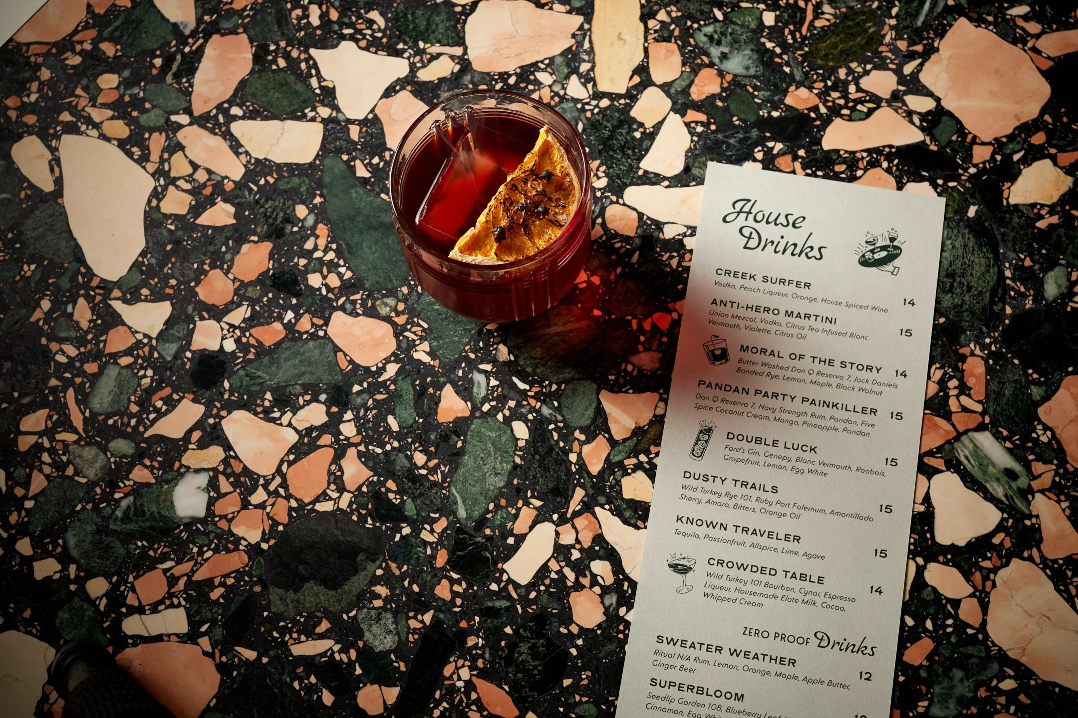 A deep red cocktail with a dried citrus slice garnish sits on a striking terrazzo surface featuring dark green, cream, and terracotta stone chips, accompanied by a "House Drinks" menu listing craft cocktails.