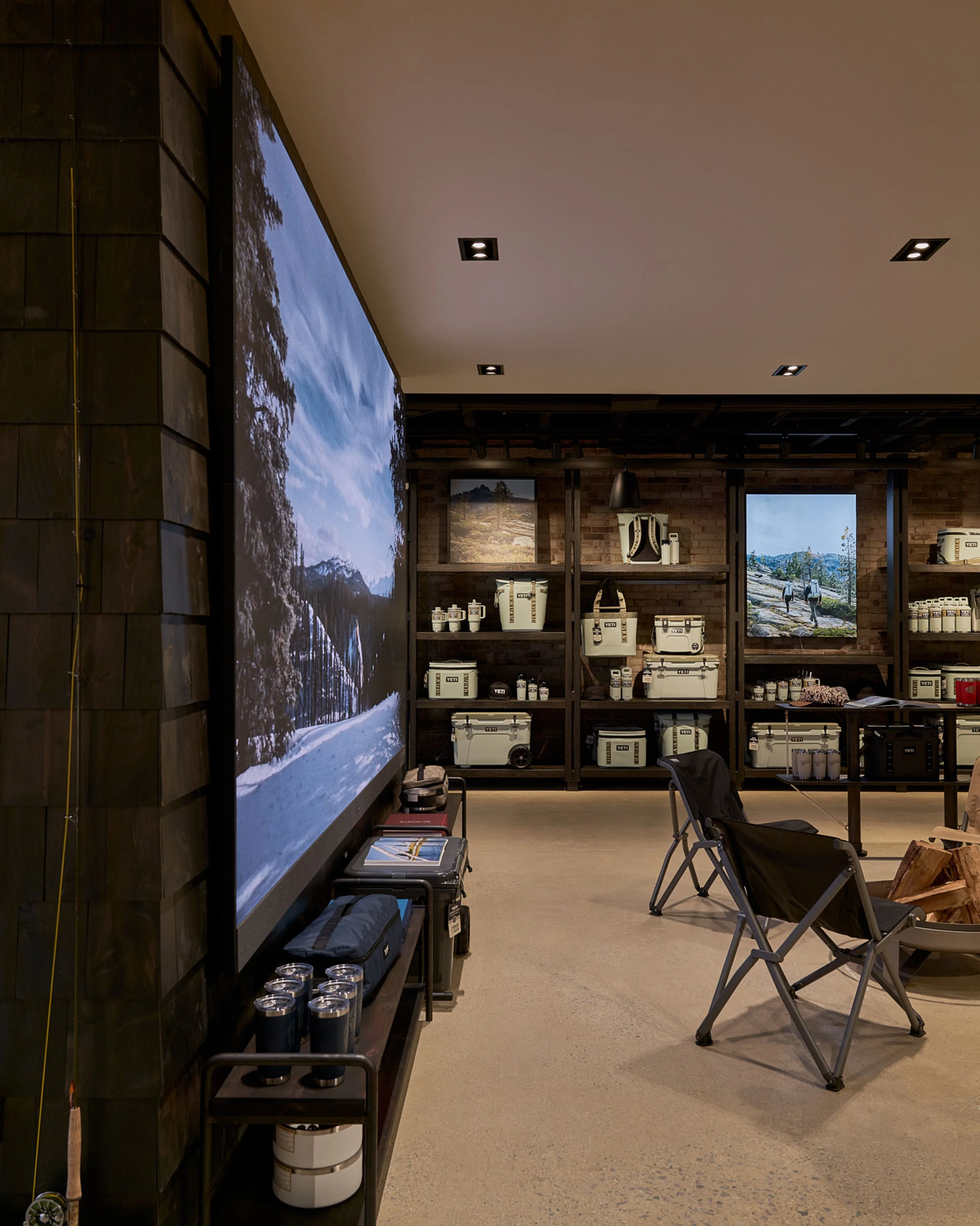 A modern retail or showroom space featuring a large video display showing a snowy mountain landscape on the left wall, with dark wood shelving units displaying cream-colored product boxes and camping gear on the back wall, minimalist black camping chairs in the foreground, and recessed ceiling lighting throughout.