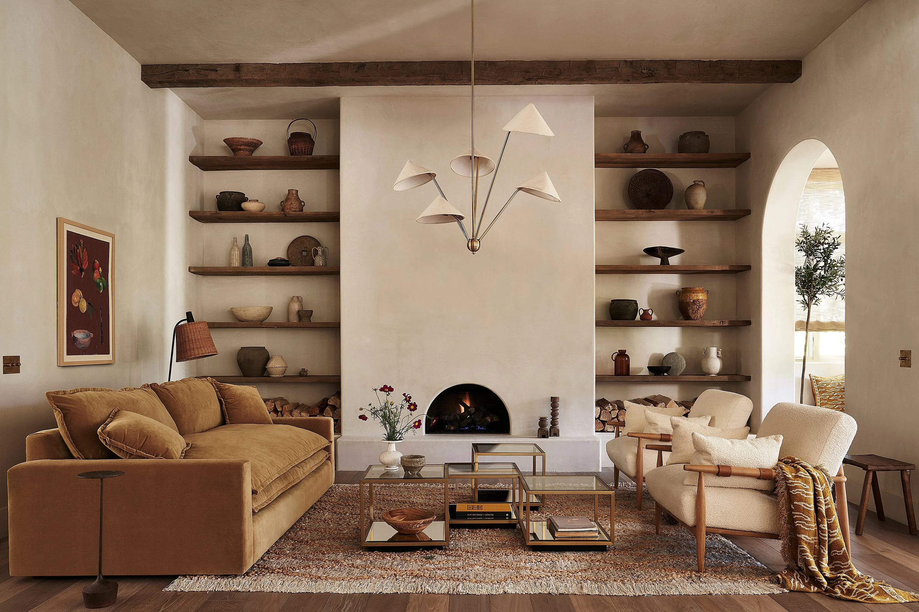 A warm, minimalist living room features a central fireplace with cream-colored plaster surround and dark wooden beam ceiling, flanked by symmetrical floating wooden shelves displaying curated ceramics and objects. The space is anchored by golden-toned upholstered seating, a sculptural pendant light fixture, and an arched doorway.