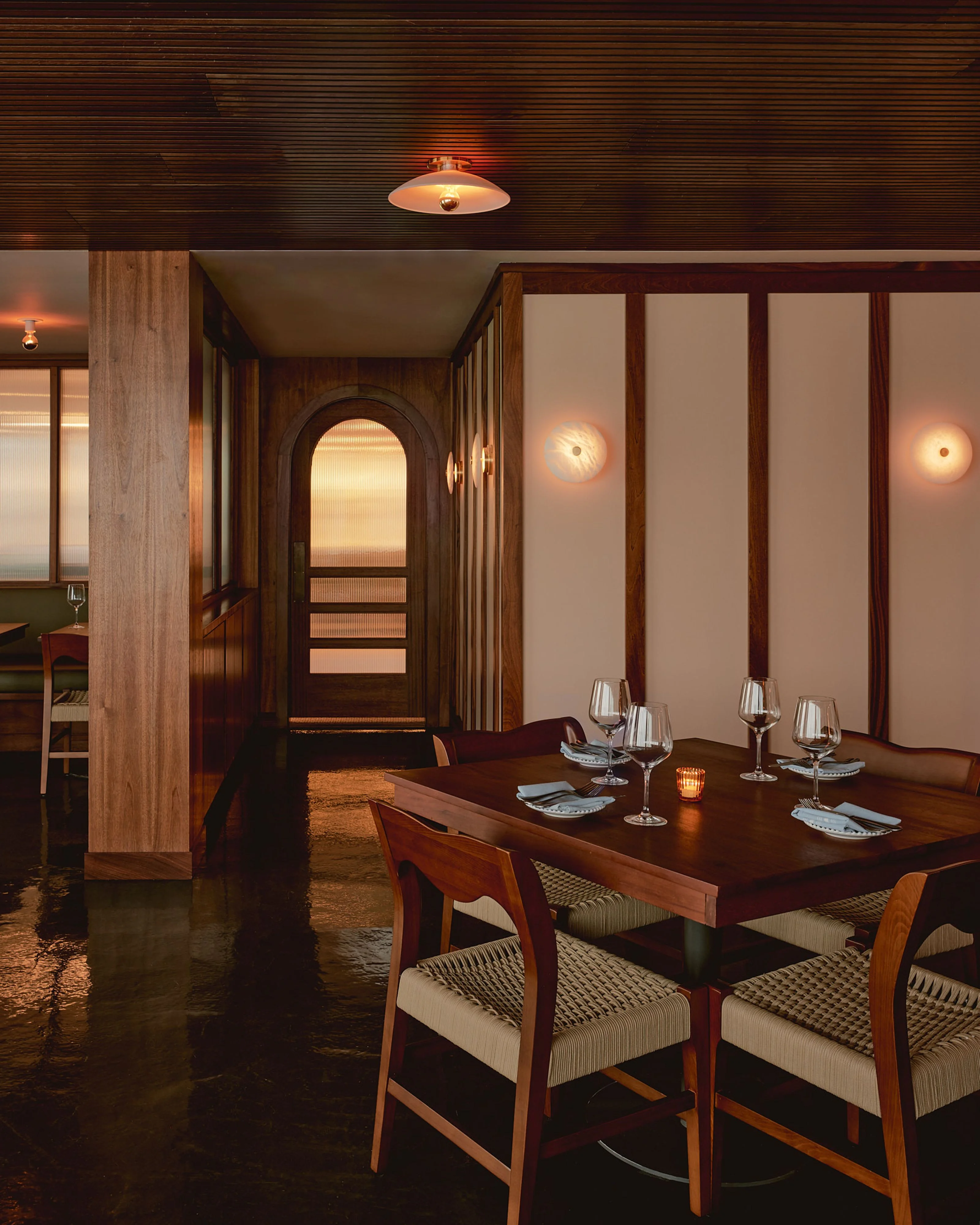 A warm, mid-century modern dining room features a dark wood table set with wine glasses and place settings, surrounded by wooden chairs with striped cream upholstery; the space is accented by rich wood paneling, cream-colored wall panels, glowing brass sconces, and polished dark flooring.