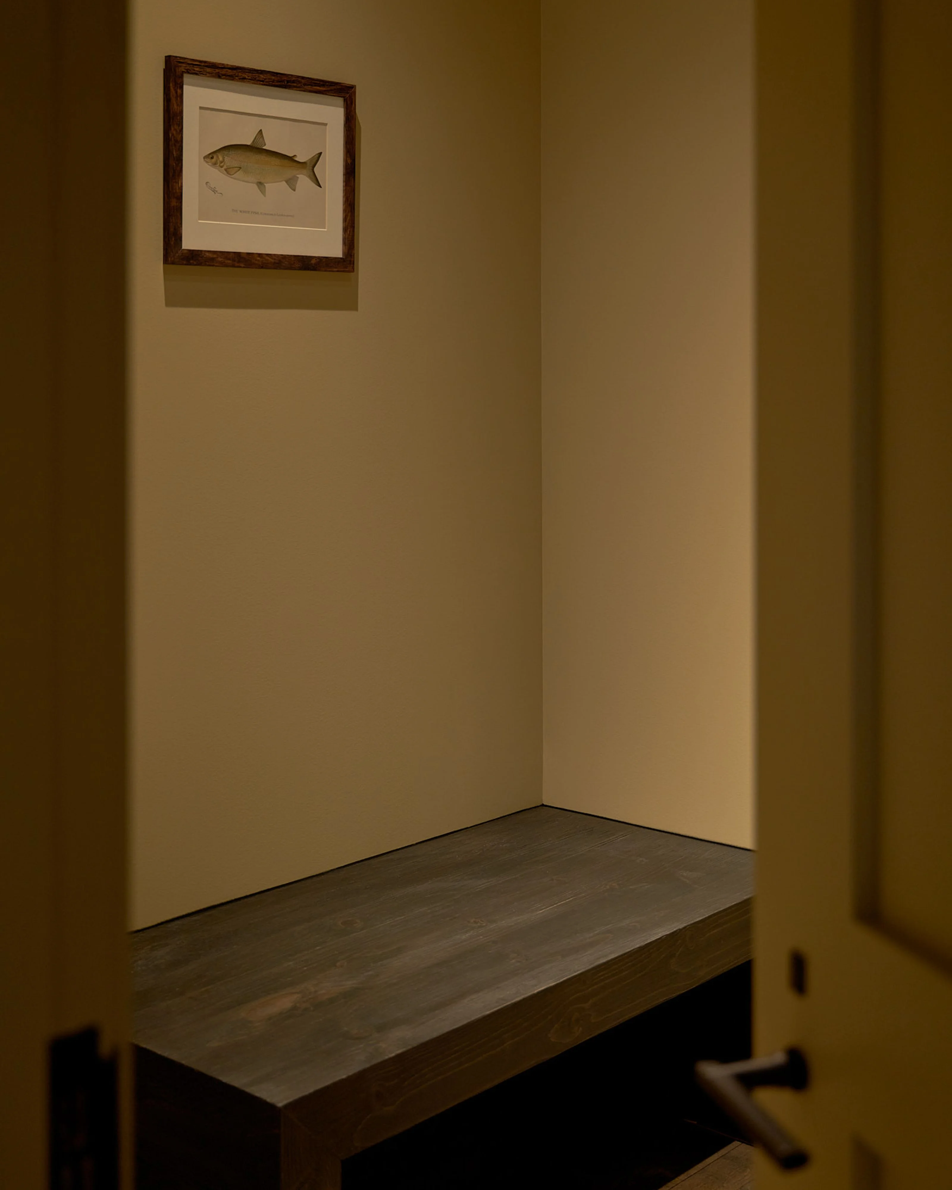 A minimalist alcove with warm beige walls features a dark wooden bench and a framed vintage fish illustration in a dark wood frame hung on the back wall, creating a serene and contemplative space with subtle warm lighting.
