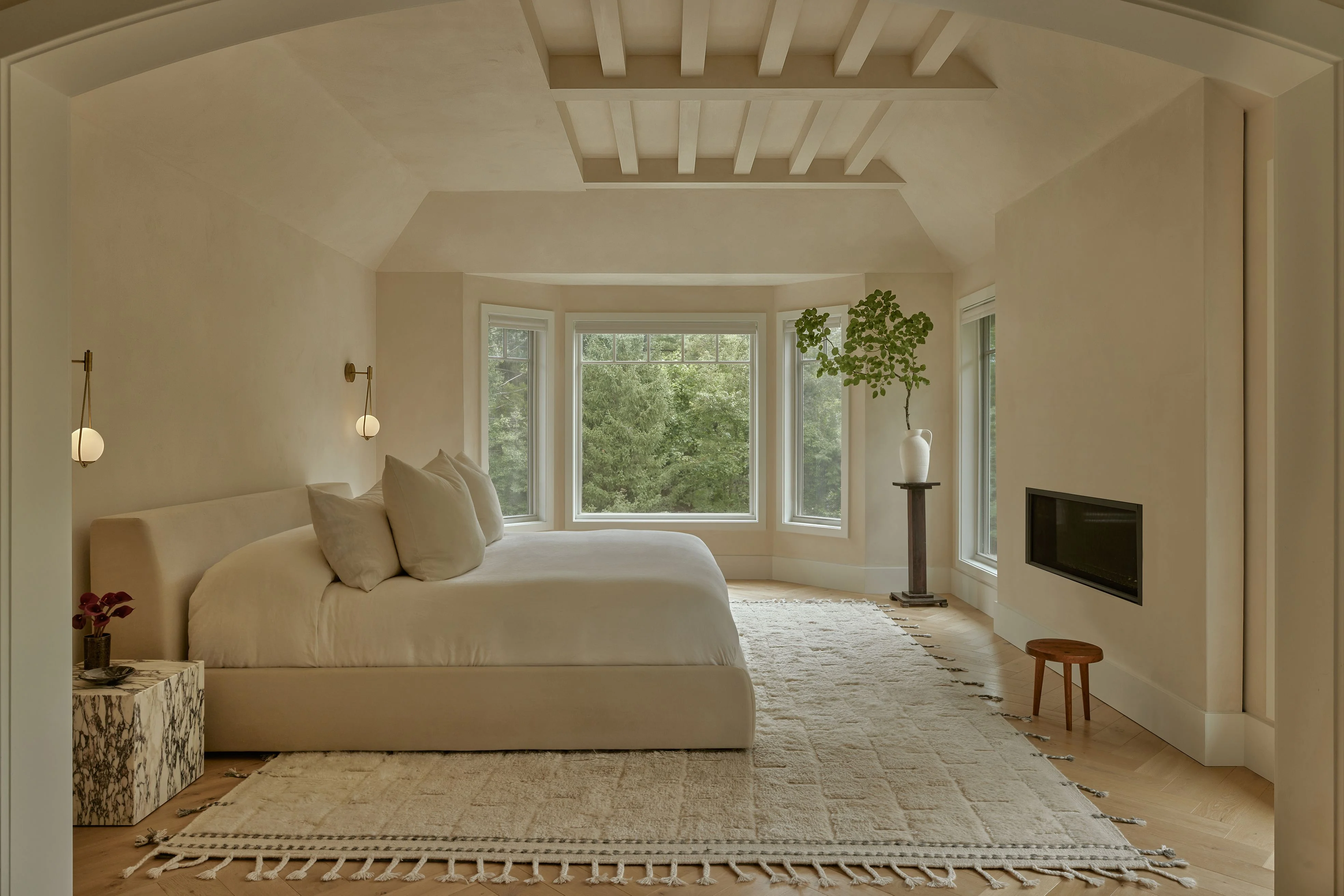 A serene, minimalist master bedroom features warm cream and beige tones throughout, with a sculptural upholstered bed positioned before expansive multi-paned windows overlooking lush greenery. The space is unified by soft neutral furnishings, exposed white ceiling beams, pendant lighting, and natural materials including woven rugs and wooden accents.