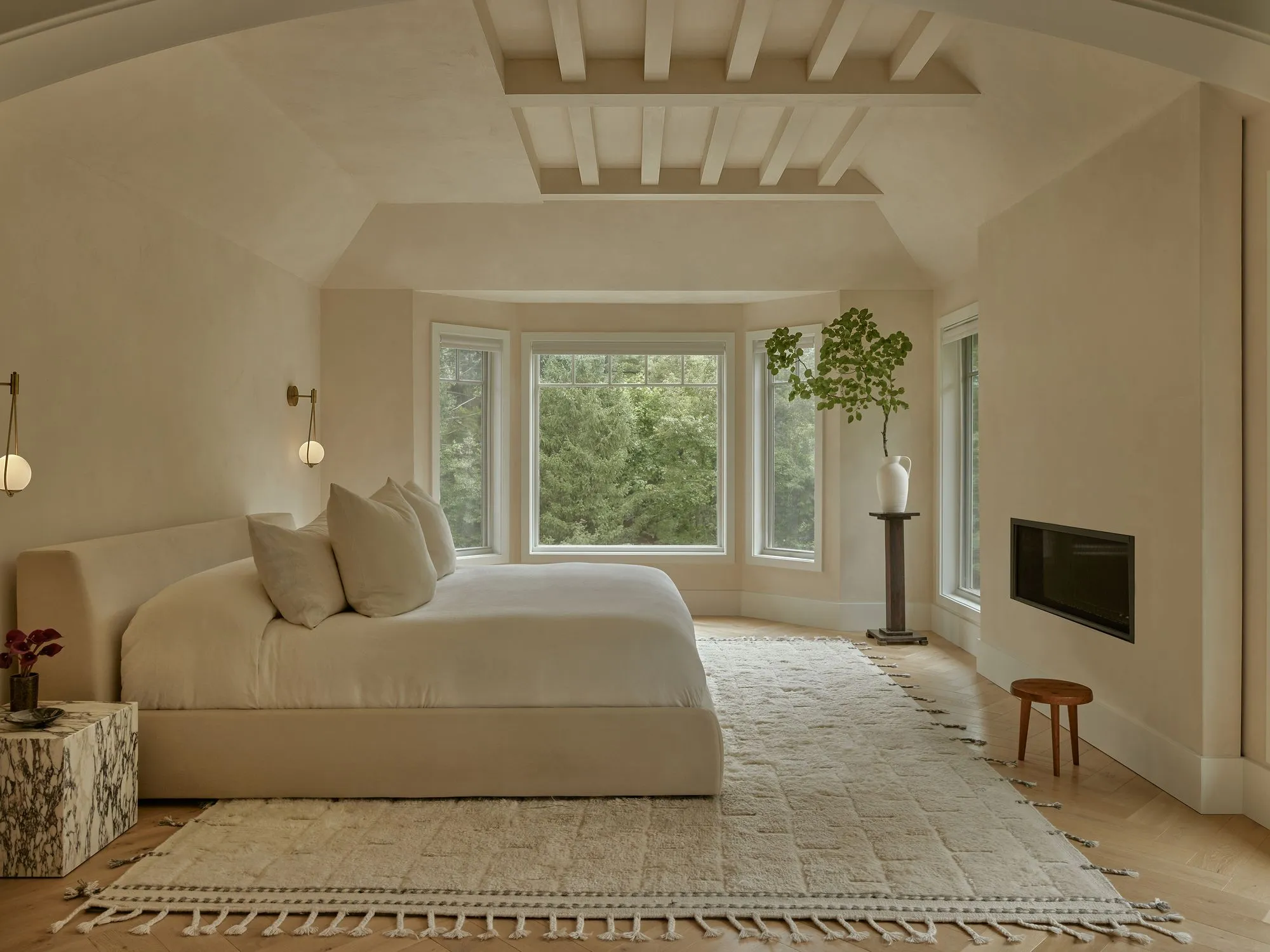 A serene, minimalist master bedroom features warm cream and beige tones throughout, with a sculptural upholstered bed positioned before expansive multi-paned windows overlooking lush greenery. The space is unified by soft neutral furnishings, exposed white ceiling beams, pendant lighting, and natural materials including woven rugs and wooden accents.