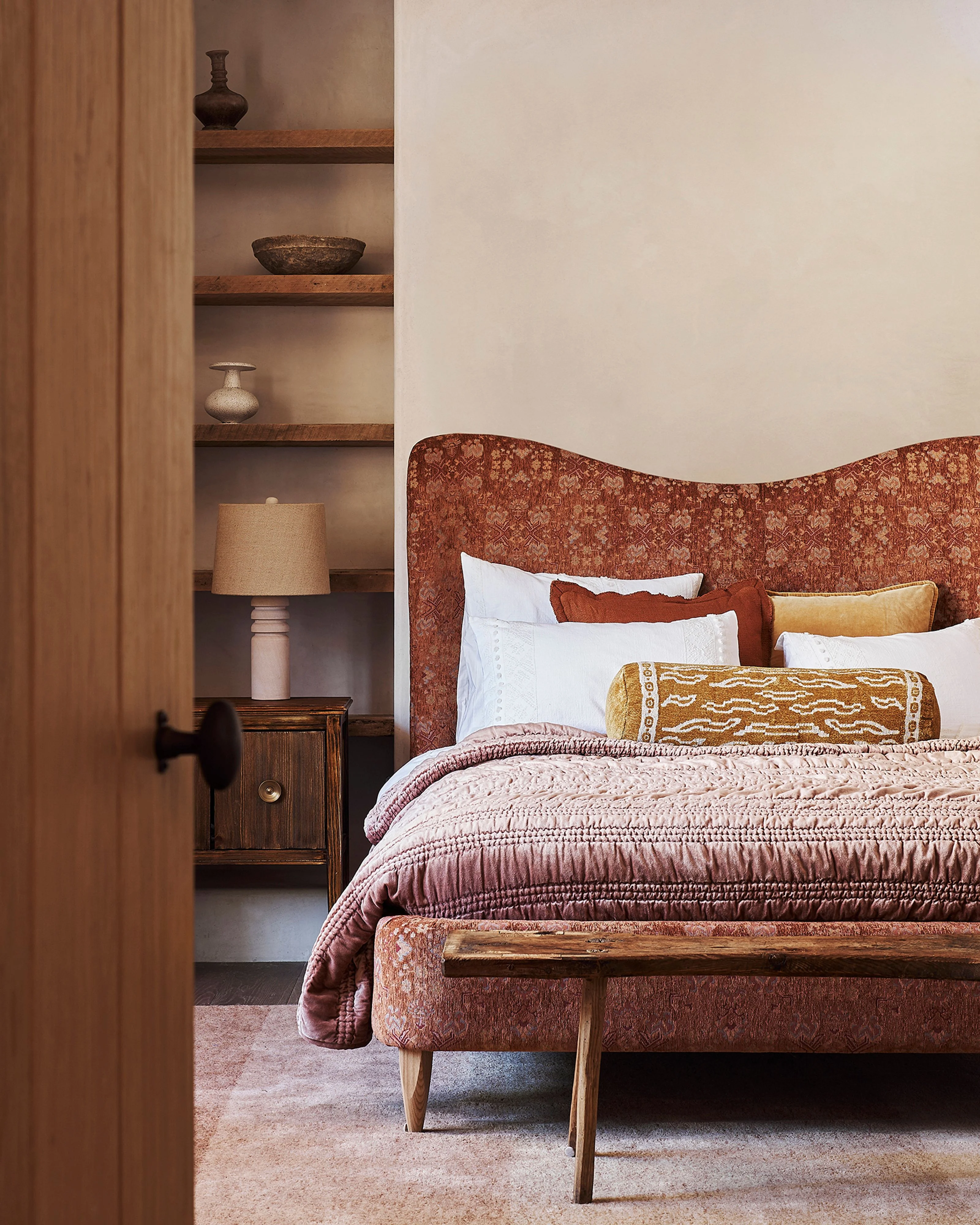 A warm, earthy-toned bedroom features a bed with a curved terracotta patterned headboard, layered with white pillows and a quilted mauve bedspread, accompanied by a matching upholstered bench at the foot. A wooden built-in shelving unit displays decorative ceramics and a table lamp, while the neutral walls and natural materials create a serene, minimalist aesthetic.