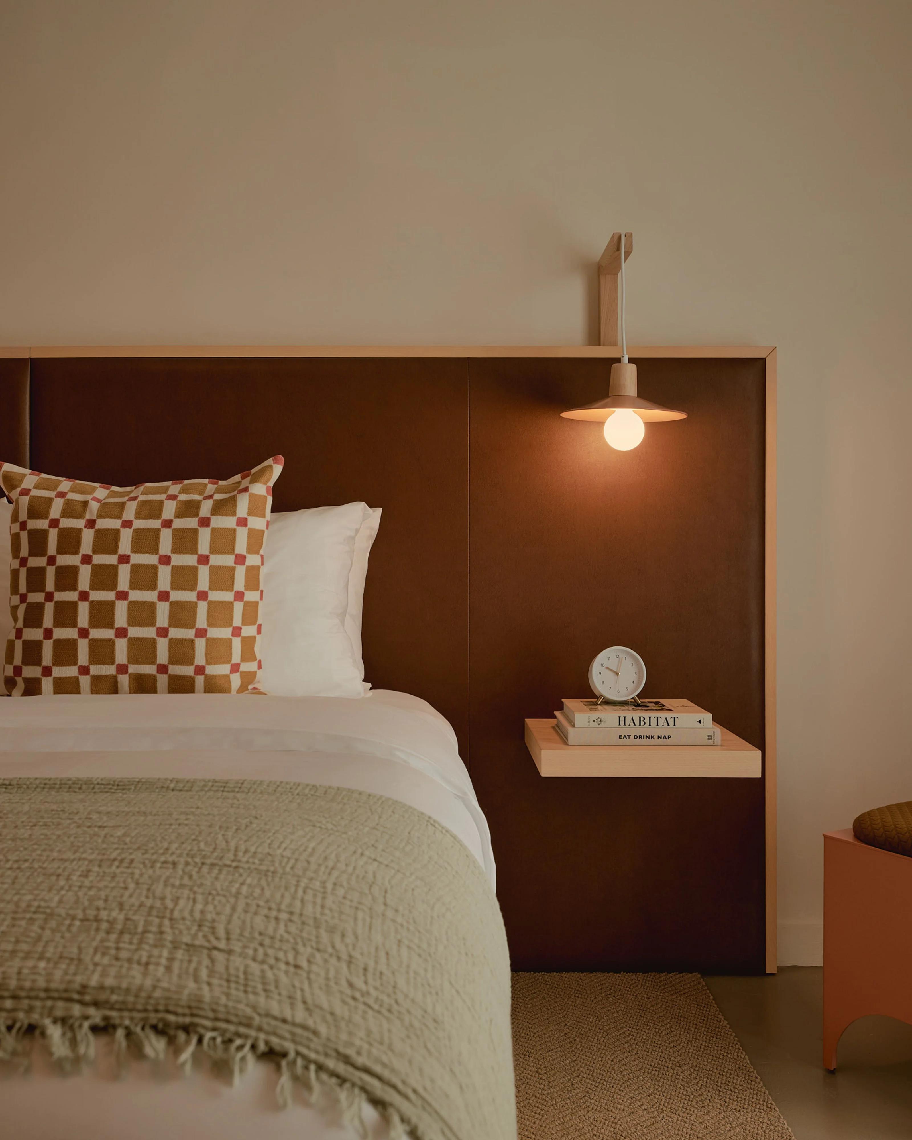 A minimalist bedroom features a bed with a checkered rust and cream pillow and sage green bedding against a tall dark brown wooden headboard with an integrated wooden nightstand. A wooden pendant light with a warm glow hangs above a small shelf displaying stacked books, a clock, and decorative items.