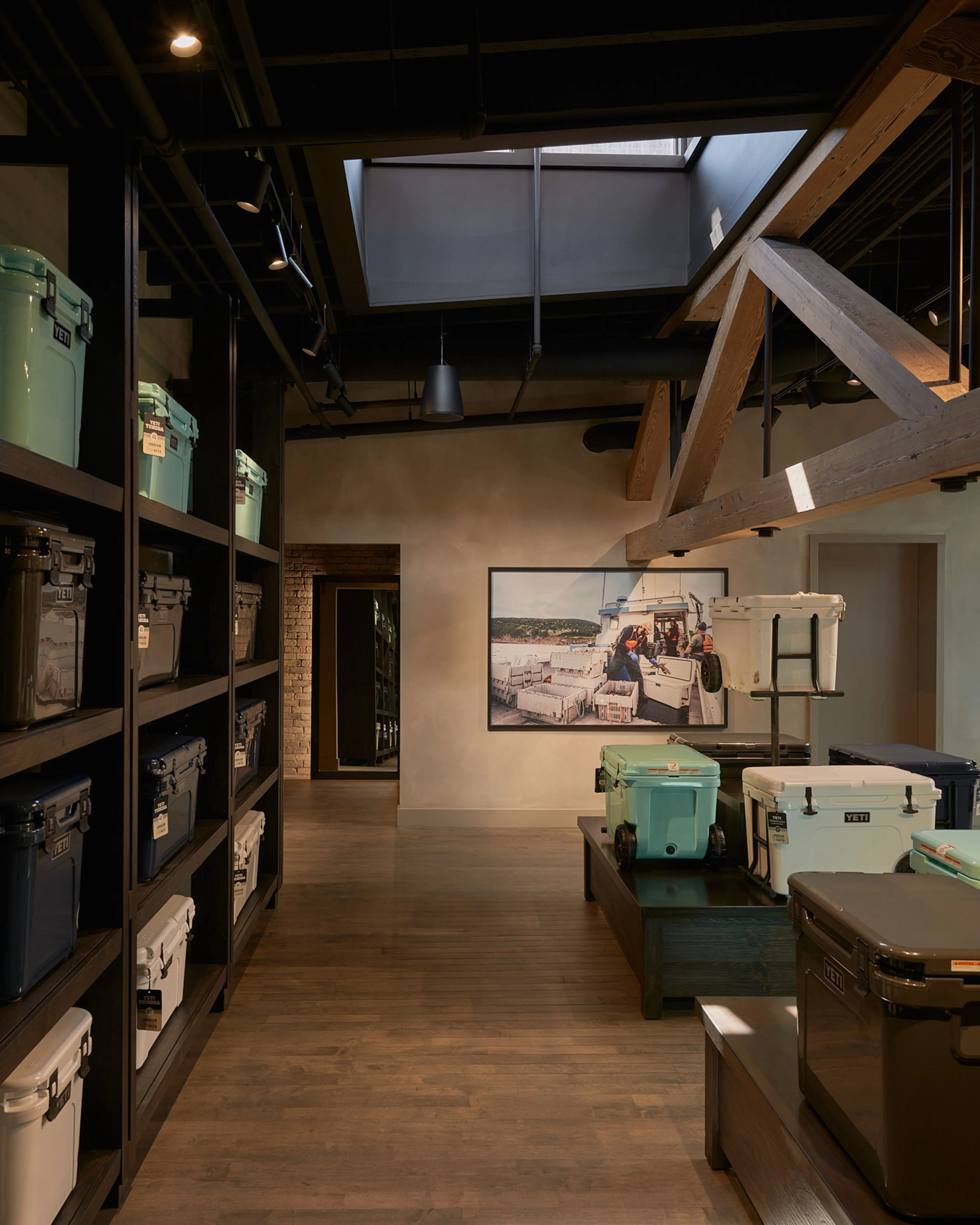 A modern industrial gallery space with exposed wooden ceiling beams and dark steel framing showcases a collection of vintage coolers and storage containers displayed on dark platforms; warm lighting illuminates cream-colored walls, built-in shelving units filled with vintage appliances, and a framed photograph of a vintage boat.