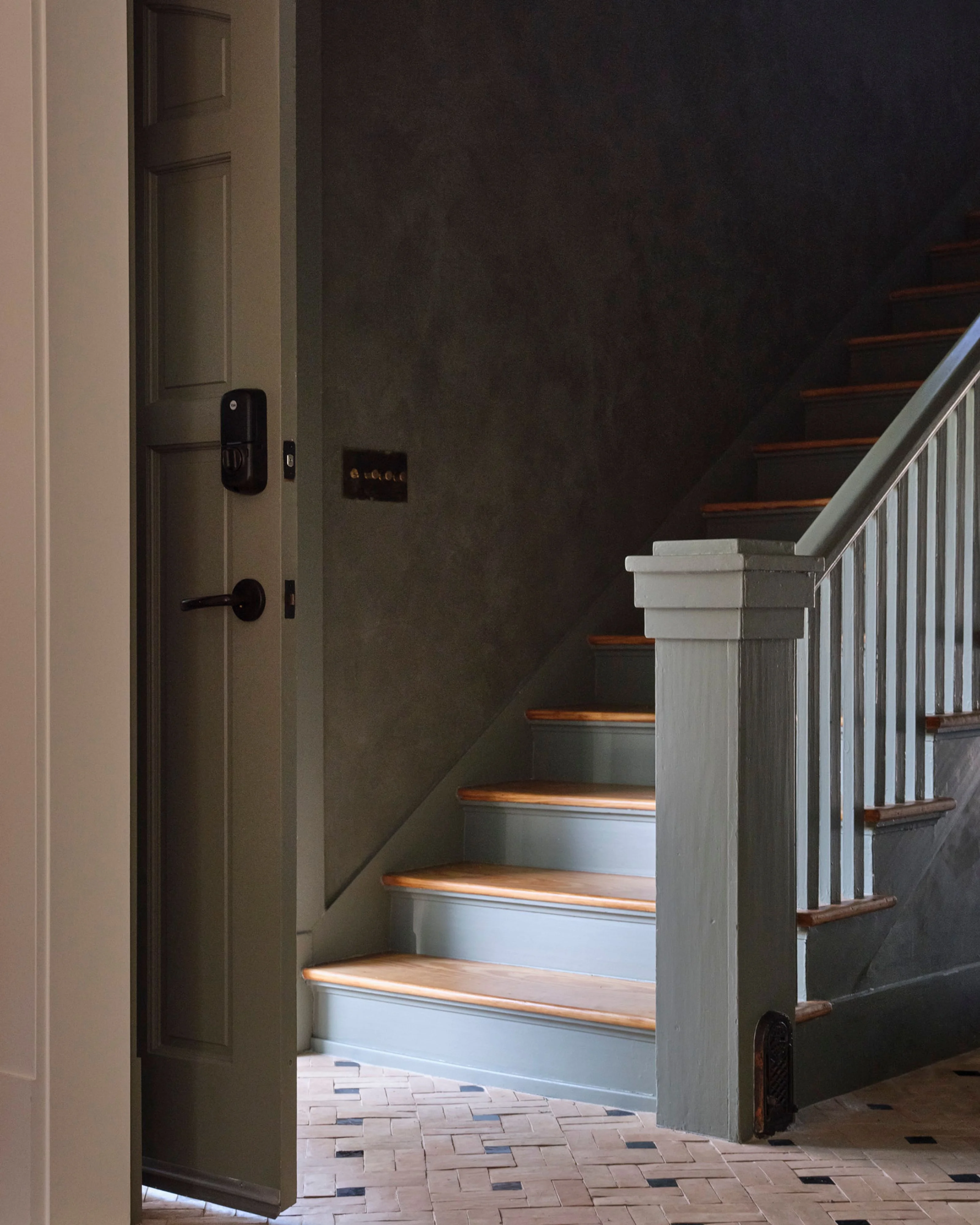 A modern staircase with warm wood treads and light blue risers descends alongside a white painted newel post and metal railing, viewed from an entryway with sage green walls and a black smart lock door. The space features a geometric patterned tile floor and clean architectural lines.