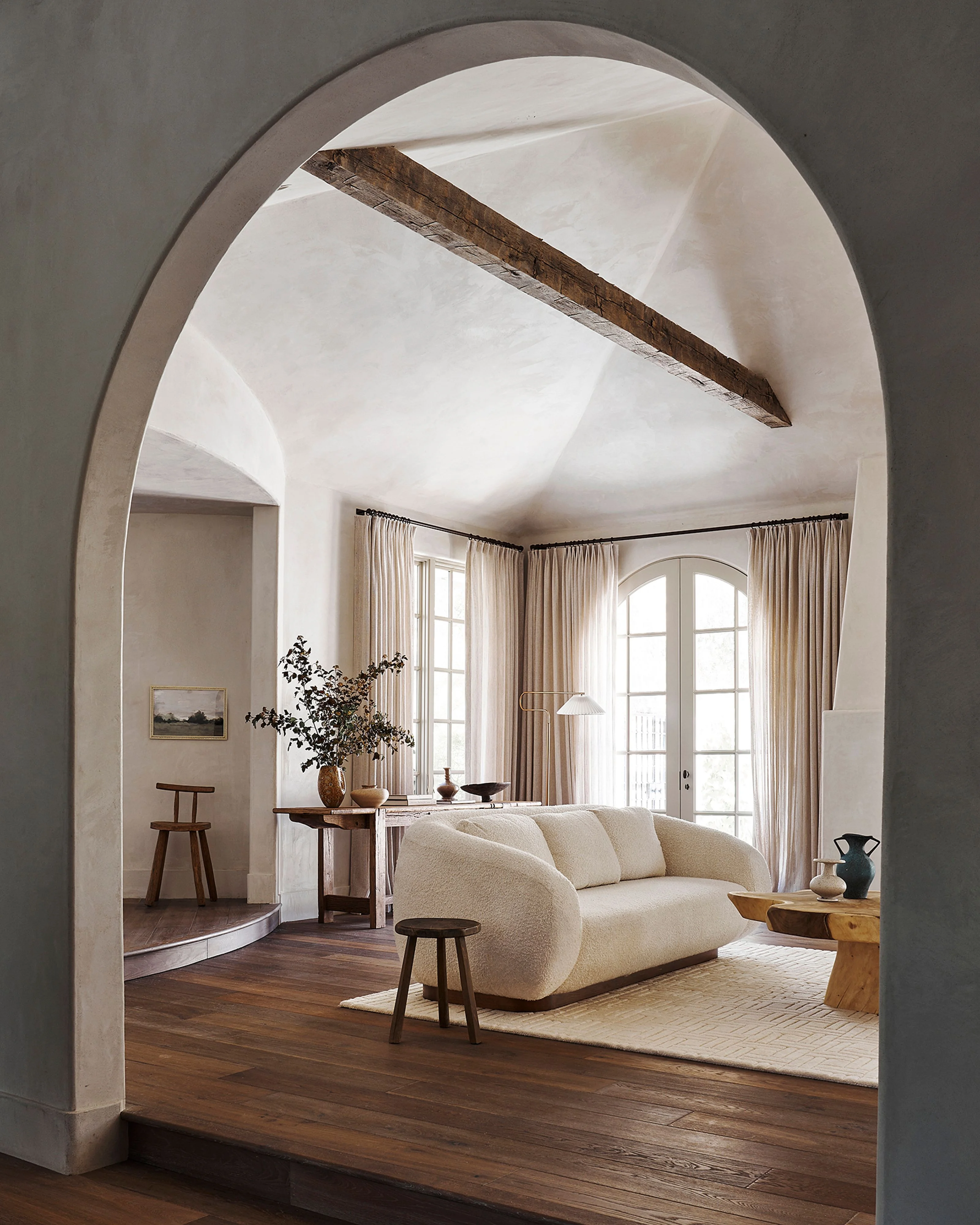A bright, minimalist living room viewed through a large rounded archway features white plaster walls, dark wooden beams, and warm wood flooring, with a cream-colored curved sofa positioned near tall arched windows dressed in neutral linen curtains that flood the space with natural light.