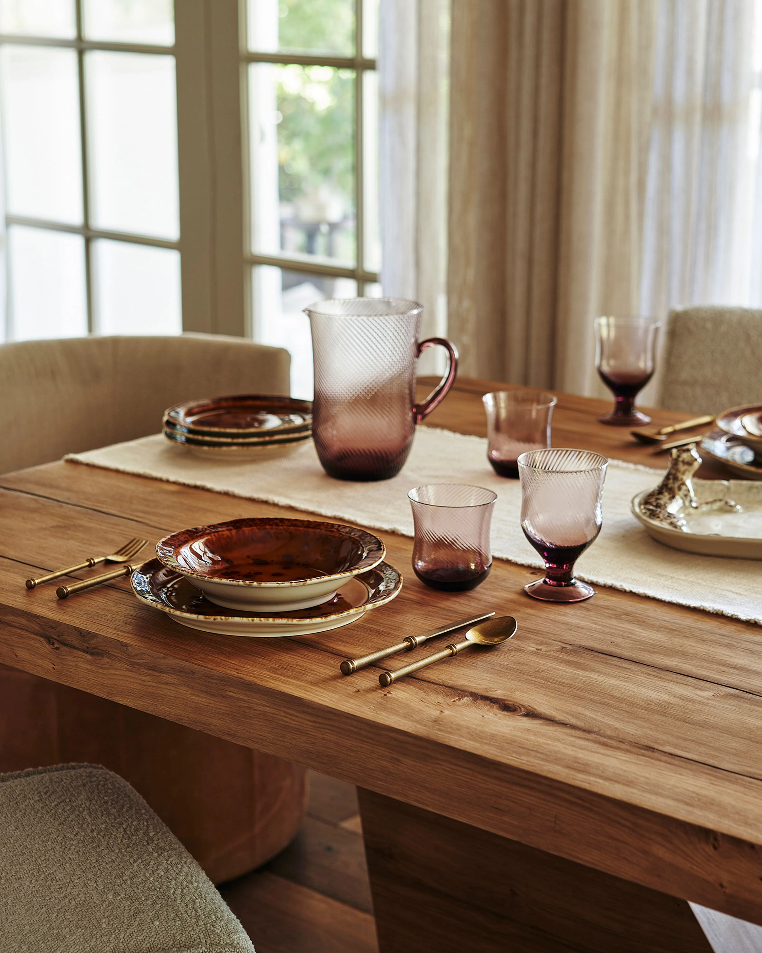 A serene dining room features a natural wood table elegantly set with burgundy and cream dinnerware, textured glassware, and brass flatware, bathed in soft natural light from large grid-pane windows draped with neutral linen curtains. Upholstered beige chairs and a cream linen table runner complete the sophisticated, minimalist aesthetic.